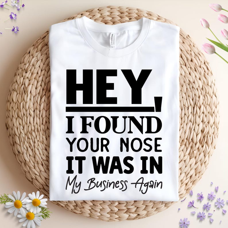 May include: A white t-shirt with the text "HEY, I FOUND YOUR NOSE IT WAS IN My Business Again" in black. The shirt is displayed on a woven wicker mat, with floral accents.