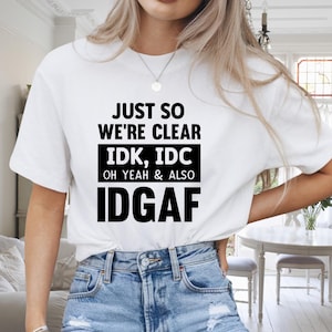 May include: White t-shirt with black text that reads "JUST SO WE'RE CLEAR IDK, IDC OH YEAH & ALSO IDGAF". The shirt is paired with blue denim jeans. The top is a casual, everyday wear item.