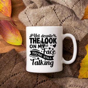 May include: White ceramic mug with a black graphic that reads "Yet despite the look on my face, you're still talking." The mug has a large handle and is set against a cozy, textured background with autumn leaves.