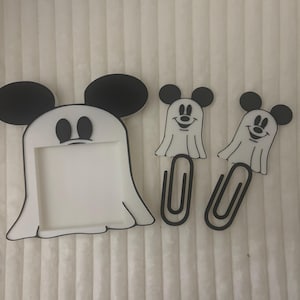 May include: A white and black Mickey Mouse ghost-shaped picture frame and two Mickey Mouse ghost paper clips. The frame has black mouse ears and a white ghost body. The paper clips have a black paper clip and a white ghost body.