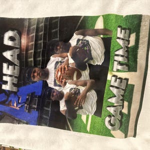 May include: White t-shirt featuring a football theme. The design includes the words "HEAD" and "GAME TIME" in bold letters, along with images of people holding a football. The background has a stadium design.