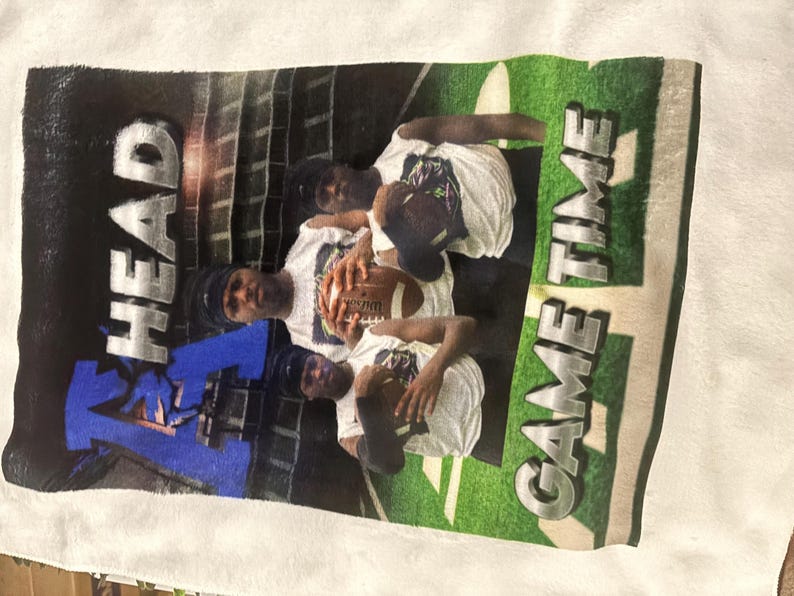 May include: White t-shirt featuring a football theme. The design includes the words "HEAD" and "GAME TIME" in bold letters, along with images of people holding a football. The background has a stadium design.