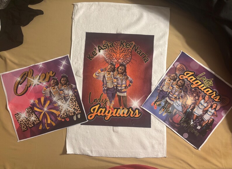 May include: Three custom printed items featuring cheerleading graphics. A white towel with a central image of two cheerleaders and the text "Lady Jaguars". Two smaller prints with cheerleaders, the word "Cheer", and the text "Ke'Asia Ke'Naria".