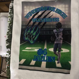 May include: White towel featuring a football field design with the text "WINDERMERE HIGH WOLVERINES" in blue and green. The towel also displays the name "JAYLEN" and the number "4". A football player is also visible.
