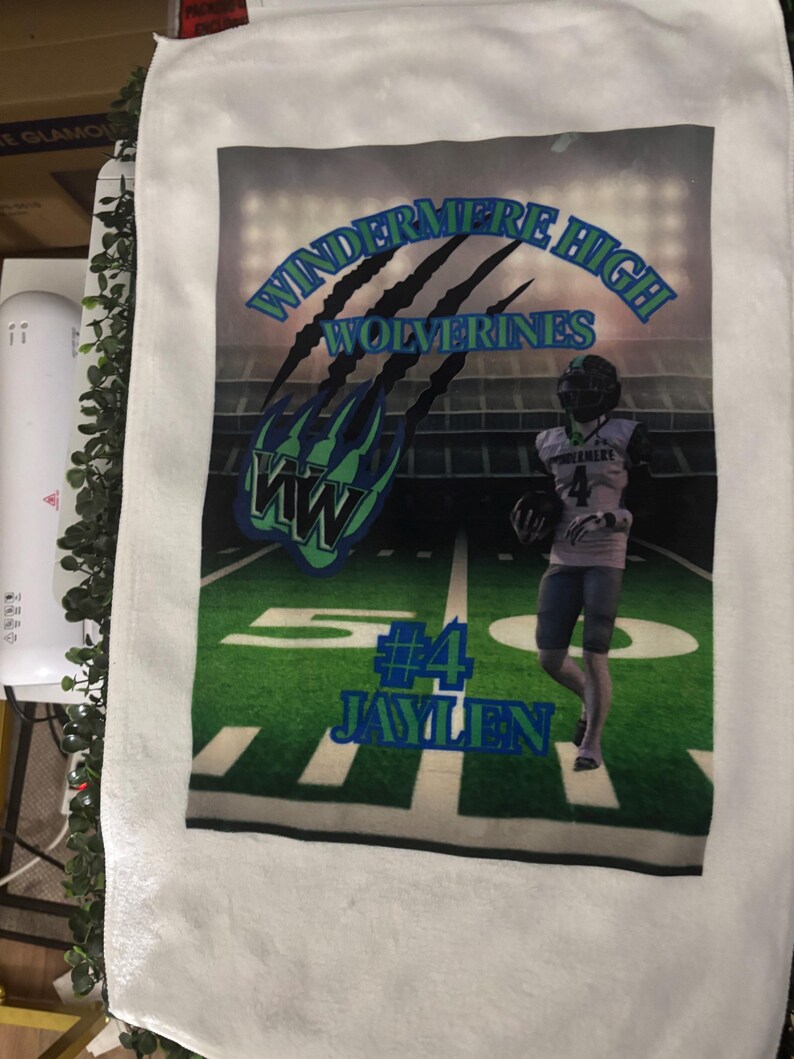 May include: White towel featuring a football field design with the text "WINDERMERE HIGH WOLVERINES" in blue and green. The towel also displays the name "JAYLEN" and the number "4". A football player is also visible.