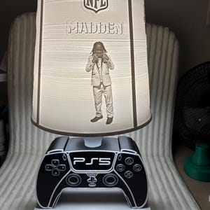 以下が含まれることがあります： 黒と白のPS5コントローラーベースと前面に「KEON」という名前が付いたカスタムランプ。 ランプシェードにはNFLのロゴ、「MADDEN」の文字、人物の画像が表示されています。