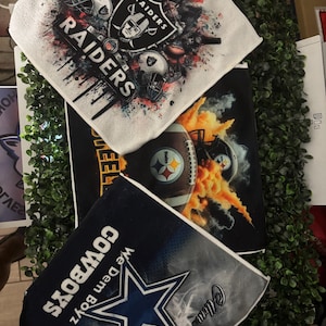 May include: Three sports-themed items: a white Raiders towel with the team logo and helmets, a black Steelers towel with a football and flames, and a navy Cowboys towel with a star and text.