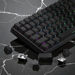 Puede incluir: Teclado mecánico negro con teclas iluminadas. El teclado tiene un diseño compacto con varias teclas de función y una tecla extraíble. Las teclas son negras con letras blancas y de colores.