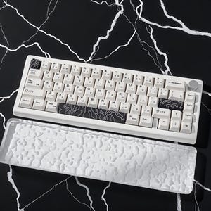 Könnte beinhalten: Eine weiße mechanische Tastatur mit japanischen Zeichen und einem schwarz-weißen topografischen Design. Die Tastatur hat einen silbernen Knopf und eine transparente, strukturierte Handballenauflage. Die Tasten sind weiß mit schwarzer Schrift.