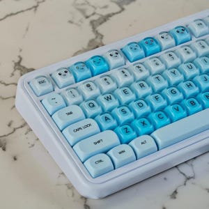 Può includere: Una tastiera meccanica con cornice bianca e tasti sfumati dal blu chiaro al blu scuro. La riga superiore dei tasti mostra varie espressioni facciali stampate, mentre i tasti rimanenti presentano lettere QWERTY nere standard.
