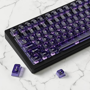 Puede incluir: Un teclado mecánico negro con teclas moradas transparentes. El teclado tiene una distribución QWERTY estándar con teclas de función y un teclado numérico. Se ven dos teclas separadas. El teclado está sobre una superficie de mármol blanco.