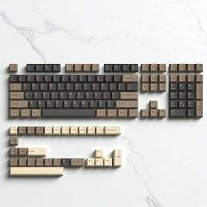 May include: A disassembled mechanical keyboard with keycaps in shades of brown, beige, and black, arranged on a white marble surface. The keycaps are organized to show the layout of the keyboard, including the main keys, number pad, and function keys.