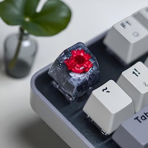 Könnte beinhalten: Eine individuelle Tastaturkappe mit einer leuchtend roten Blume, die in einem transparenten, harzähnlichen Material eingeschlossen ist. Die Kappe befindet sich auf einer grauen Tastatur mit weißen Tasten. Die Details der Blume sind sichtbar.