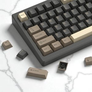 May include: A close-up of a mechanical keyboard with dark gray keys and beige and brown accent keys. The keyboard has a dark gray frame. Several keycaps are detached and scattered around the keyboard.
