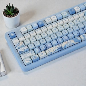Puede incluir: Un teclado mecánico azul claro con teclas personalizadas. Las teclas presentan diseños azules y blancos, incluyendo criaturas marinas y patrones. El teclado tiene un marco azul claro y una pequeña planta suculenta en una maceta blanca.