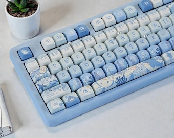 Puede incluir: Un teclado mecánico azul claro con teclas personalizadas. Las teclas presentan diseños azules y blancos, incluyendo criaturas marinas y patrones. El teclado tiene un marco azul claro y una pequeña planta suculenta en una maceta blanca.