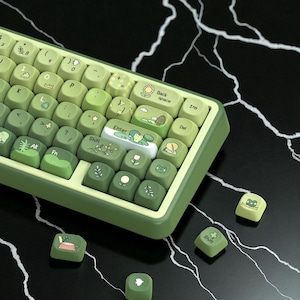 Puede incluir: Un teclado mecánico verde y blanco con teclas personalizadas. Las teclas presentan varias ilustraciones de plantas, ranas y otros diseños con temas de la naturaleza. El teclado tiene un esquema de color degradado, que va del verde claro al oscuro.