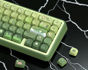 May include: A green and white mechanical keyboard with custom keycaps. The keycaps feature illustrations of plants, frogs, and nature-themed designs. The keyboard has a gradient colour scheme, transitioning from light to dark green.