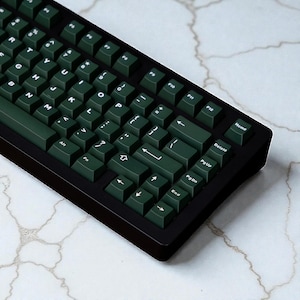 May include: A black mechanical keyboard with dark green keycaps. The keyboard has a standard layout with letters, numbers, and function keys. The keycaps are a solid, dark green color with white lettering. The keyboard is angled slightly towards the user.