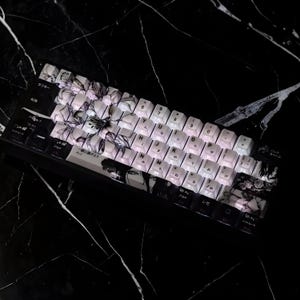 Może przedstawiać: Czarna klawiatura mechaniczna z białymi nakładkami na klawisze z grafiką w stylu anime. Klawisze wyświetlają japońskie znaki i angielskie litery. Klawiatura leży na czarno-białej marmurowej powierzchni.