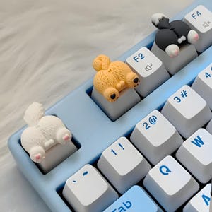 Puede incluir: Un teclado azul claro con teclas blancas y tres teclas con forma de perro. Las teclas tienen forma de trasero de perro, con una combinación de colores blanco, beige y negro. El teclado está sobre una superficie blanca y esponjosa.