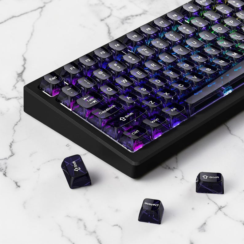 May include: Black mechanical keyboard with transparent keycaps. The keys are backlit with a rainbow of colours. Three detached keycaps are scattered nearby. The keyboard has a black frame and a compact design.