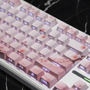 Puede incluir: Un teclado mecánico blanco con teclas rosas y blancas. Las teclas presentan diseños de flores de cerezo y patrones de degradado rosa. El teclado tiene un diseño compacto con teclas de función y una prominente tecla Enter.
