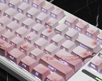 May include: A white mechanical keyboard with pink and white keycaps. The keycaps feature cherry blossom designs and pink gradient patterns. The keyboard has a compact layout with function keys and a prominent Enter key.