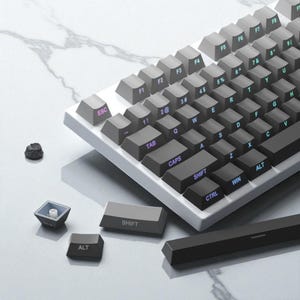 Może przedstawiać: Klawiatura mechaniczna z szarymi klawiszami i białą ramką. Klawisze są oznaczone białymi i niebieskimi literami. Kilka klawiszy jest odłączonych, odsłaniając elementy wewnętrzne. W pobliżu leży mała, ciemna skała.