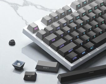 Op de afbeelding: Een mechanisch toetsenbord met grijze toetsen en een wit frame. De toetsen zijn gemarkeerd met witte en blauwe letters. Verschillende toetsen zijn losgemaakt, waardoor de interne componenten zichtbaar zijn. Een kleine, donkere steen ligt in de buurt.