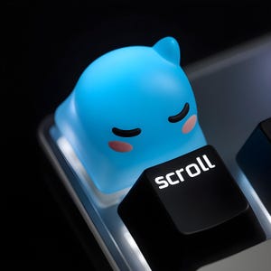May include: A light blue, cartoon-style keycap shaped like a sleeping creature, with black eyes and pink cheeks, sits atop a keyboard. The black key below it reads "scroll" in white lettering. The keycap is a unique accessory.
