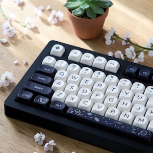 Puede incluir: Un teclado mecánico en blanco y negro con un diseño espacial. El teclado presenta teclas blancas con letras negras y una barra espaciadora azul oscuro. Una pequeña planta suculenta en maceta y flores blancas en el fondo.