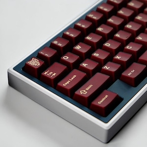 Könnte beinhalten: Nahaufnahme einer mechanischen Tastatur mit weinroten Tastenkappen und weißer Beschriftung. Die Tastatur hat einen silbernen Rahmen und eine dunkelblaue Basis. Die Tasten sind in einem Standard-QWERTZ-Layout angeordnet.