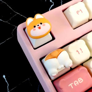 Pode incluir: Uma imagem em close-up de um teclado mecânico rosa com teclas personalizadas. Uma tecla é um rosto de cachorro Shiba Inu de desenho animado, e outra é a parte traseira do cachorro. Outras teclas são brancas e rosas.