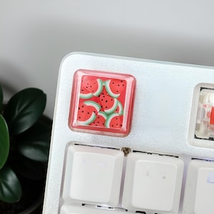 May include: A square, red and clear keyboard keycap with a watermelon design. The keycap features a pattern of watermelon slices with red rinds, green edges, and black seeds. The keycap is set on a white keyboard.