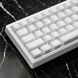 May include: A close-up shot of a white mechanical keyboard with white keycaps. The keyboard has a compact design, with a minimalist aesthetic. The keycaps are uniformly colored and feature clear, printed characters. The keyboard is set against a dark, marbled surface.