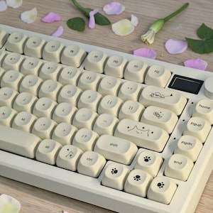 Puede incluir: Un teclado mecánico de color crema con teclas personalizadas. Las teclas presentan varios diseños con temática de gatos y huellas de patas. El teclado tiene un marco blanco y una pequeña pantalla. Pétalos y vegetación están esparcidos alrededor del teclado.