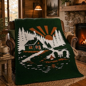 May include: A dark green crocheted blanket featuring a cabin scene with a rising sun, trees, and a lake. The design is in white and orange, creating a cozy, rustic aesthetic. The blanket is draped over a leather armchair.