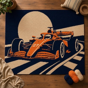 May include: A rectangular crochet rug featuring a dark navy blue background with a large beige circular moon. An orange race car with the number 27 on its nose is depicted driving along a beige track with white stripes against the dark backdrop.