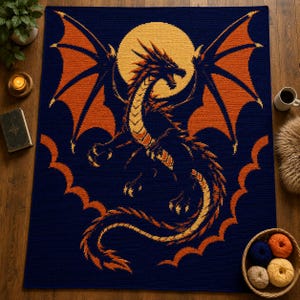 Puede incluir: Una manta de ganchillo rectangular con un fondo azul marino oscuro y una gran luna amarilla central. Un dragón detallado con alas y cuerpo de color naranja y amarillo está centrado frente a la luna, con detalles decorativos de borde naranja.