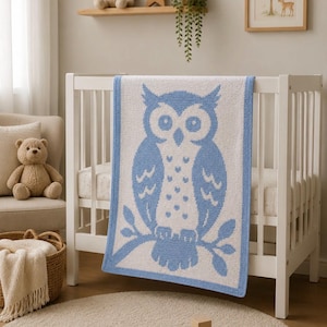 May include: A blue and white owl-patterned baby blanket is draped over a white cot. The owl design features a detailed illustration of the bird. The blanket is made of soft material, suitable for a nursery, and is a decorative item.