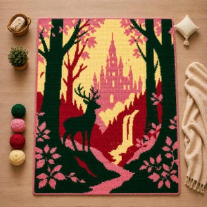 May include: A rectangular crochet blanket featuring a forest scene with a dark green deer silhouette, a pink castle, and a yellow waterfall. The design is framed by dark green trees with pink leaves, all bordered by a thin pink edge on a light background.