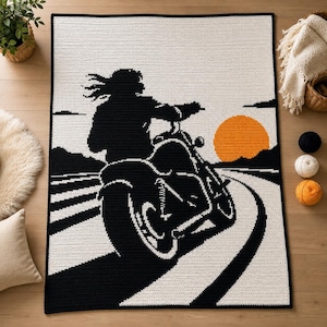 Puede incluir: Una manta de ganchillo rectangular que presenta la silueta negra de una persona montando una motocicleta en una carretera. El diseño incluye un gran sol naranja sobre un fondo blanco, con líneas horizontales negras que representan la carretera.