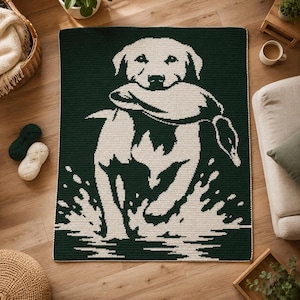 May include: A rectangular crochet blanket featuring a dark forest green background with a cream-colored graphic design of a dog carrying a duck in its mouth while splashing through water. The blanket has a simple, clean border and flat texture.