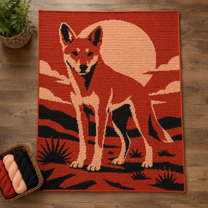 May include: A rectangular crochet blanket featuring a graphic design of a dingo standing in a desert landscape at sunset. The scene includes a large pale orange sun, dark black silhouettes of desert plants, and a vibrant rust-colored background.