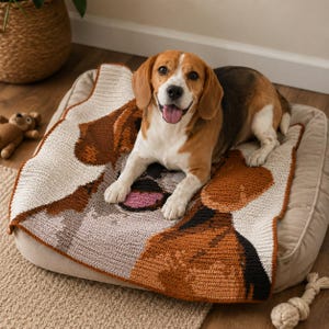 Pode incluir: Um cão beagle está deitado numa cama para cães bege, sobre uma manta de crochê com um desenho de rosto de beagle em tons de castanho, branco e cinza. Um pequeno urso de pelúcia e um brinquedo de corda são visíveis. O cão está sorrindo com a língua de fora.