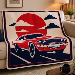 May include: A crocheted blanket featuring a red car design set against a red sun and blue clouds. The blanket has a cream-colored background with a navy blue border. The car is detailed with navy blue accents, creating a retro aesthetic.