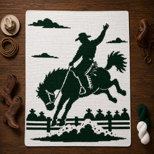 May include: A rectangular crochet blanket featuring a dark green graphic design on a white background. The pattern depicts a cowboy riding a bucking horse, jumping over a fence, with clouds in the sky. The blanket has a textured, woven appearance.