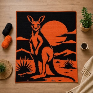 May include: A rectangular crochet blanket featuring a high-contrast design in black and orange. The pattern depicts a kangaroo standing in a desert landscape with spiky plants and a large, circular sun against a dark sky background.