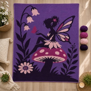 May include: A rectangular crochet blanket featuring a purple background with a dark purple silhouette of a fairy sitting on a spotted mushroom. The design includes light pink flowers, a small butterfly, and a flower at the base in shades of pink.